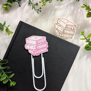 May include: A white paperclip bookmark featuring a pink stack of books design. The bookmark is on a black notebook, with a second stack of books design in brown. Greenery and white faux fur are in the background.