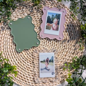 Puede incluir: Tres marcos de fotos con bordes ondulados en acrílico verde, rosa y transparente, cada uno con pequeños cierres plateados. Dos marcos contienen fotos instantáneas de personas. Los marcos descansan sobre una estera tejida, rodeados de vegetación.