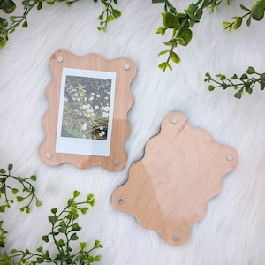 May include: Two wooden photo frames with a wavy edge design. One frame holds a polaroid photo of white flowers. The frames are light brown with clear acrylic covers and small magnets to hold the photo in place.