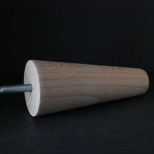 May include: A wooden furniture leg with a tapered design and a metal screw. The leg is light brown and has a smooth finish.