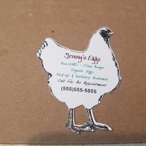 May include: A white chicken-shaped sticker with black outlines and legs. The sticker reads "Jenny's Eggs" with text indicating non-GMO, free-range, and organic eggs. It also mentions pick-up and delivery availability and a phone number.