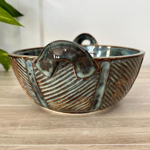 May include: A handcrafted ceramic bowl with a unique design. The bowl features a textured exterior with a ribbed pattern and a handle. The color is a blend of teal, brown, and blue, giving it a rustic and artistic appearance. The bowl is suitable for serving or display.