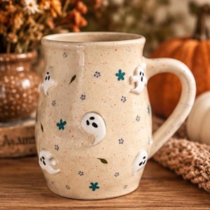May include: A cream-colored ceramic mug with a speckled finish, featuring raised white ghost figures and small blue floral accents. The mug has a rounded body, a sturdy handle, and a smooth glazed surface, showcasing a repeating pattern of ghosts.