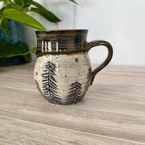 Könnte beinhalten: Eine handgefertigte Keramik-Tasse mit dunkelbraunem Rand und Henkel. Der Korpus der Tasse ist cremefarben mit einem schwarzen Baumdesign und kleinen schwarzen Punkten. Die Tasse steht auf einer hellen Holzoberfläche.