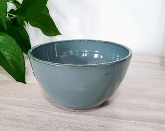 May include: A ceramic bowl with a matte, dusty blue exterior. The bowl has a rounded shape with a slightly flared rim. The interior is a lighter shade of blue. The bowl is sitting on a light wood surface.