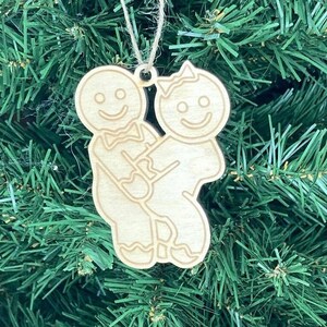 May include: A wooden Christmas ornament featuring two gingerbread people holding hands. The ornament is a light brown color and has a simple design.