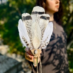 Puede incluir: Un abanico artesanal de plumas blancas, grises y negras. El abanico presenta un mango de cuero marrón con un cristal transparente y una pequeña concha. Las plumas están dispuestas en forma de abanico, con una estética natural y terrosa.