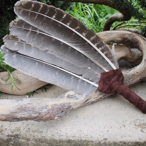 May include: A brown leather-wrapped feather smudge stick with a fringe. The feathers are gray and white and are arranged in a fan shape.