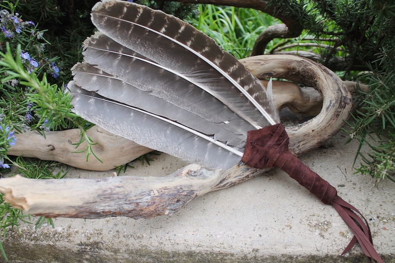 May include: A brown leather-wrapped feather smudge stick with a fringe. The feathers are gray and white and are arranged in a fan shape.