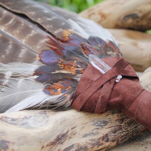 May include: A brown leather-wrapped smudge stick with a clear quartz crystal at the end. The smudge stick is made of feathers and is resting on a piece of driftwood.