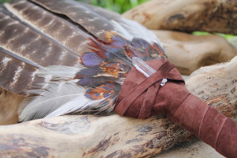 May include: A brown leather-wrapped smudge stick with a clear quartz crystal at the end. The smudge stick is made of feathers and is resting on a piece of driftwood.