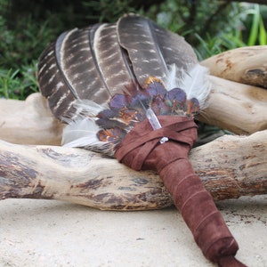 May include: A brown leather-wrapped feather fan with a clear crystal at the center. The fan is resting on a piece of driftwood.