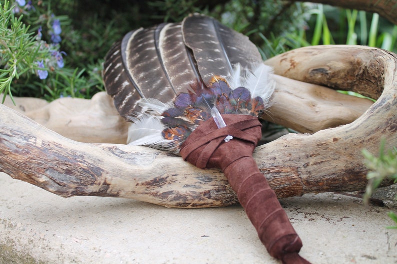 May include: A brown leather-wrapped feather fan with a clear crystal at the center. The fan is resting on a piece of driftwood.