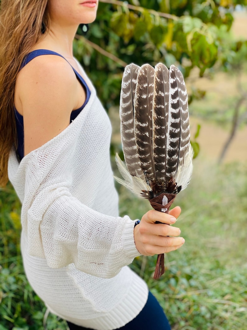 May include: A white feather smudge stick with brown leather ties. The feathers are arranged in a fan shape and have black and white markings. The smudge stick is held by a person wearing a white sweater.