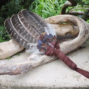 May include: A brown leather-wrapped feather fan with a clear crystal at the center. The fan is resting on a piece of driftwood.