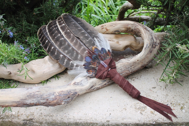 May include: A brown leather-wrapped feather fan with a clear crystal at the center. The fan is resting on a piece of driftwood.