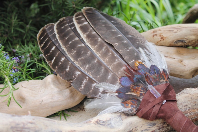May include: A brown and white feather fan with blue, red, and orange accents. The fan is tied with a brown leather strap and has a clear crystal attached.