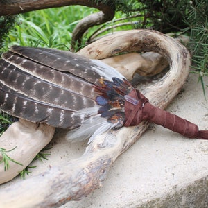May include: A brown leather-wrapped feather fan with a long brown tassel. The fan is made of various feathers, including brown, gray, and white feathers. The fan is lying on a piece of driftwood.