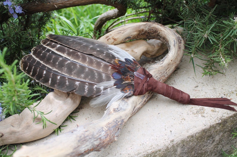 May include: A brown leather-wrapped feather fan with a long brown tassel. The fan is made of various feathers, including brown, gray, and white feathers. The fan is lying on a piece of driftwood.