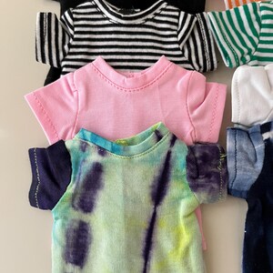 Puede incluir: Una colecci&oacute;n de camisetas peque&ntilde;as para mu&ntilde;ecas o juguetes, con varios dise&ntilde;os que incluyen una camiseta negra lisa, una camiseta a rayas blancas y negras, una camiseta rosa lisa, una camiseta tie-dye morada y verde, y otros patrones de rayas coloridas.