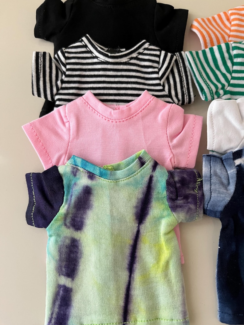 Puede incluir: Una colecci&oacute;n de camisetas peque&ntilde;as para mu&ntilde;ecas o juguetes, con varios dise&ntilde;os que incluyen una camiseta negra lisa, una camiseta a rayas blancas y negras, una camiseta rosa lisa, una camiseta tie-dye morada y verde, y otros patrones de rayas coloridas.