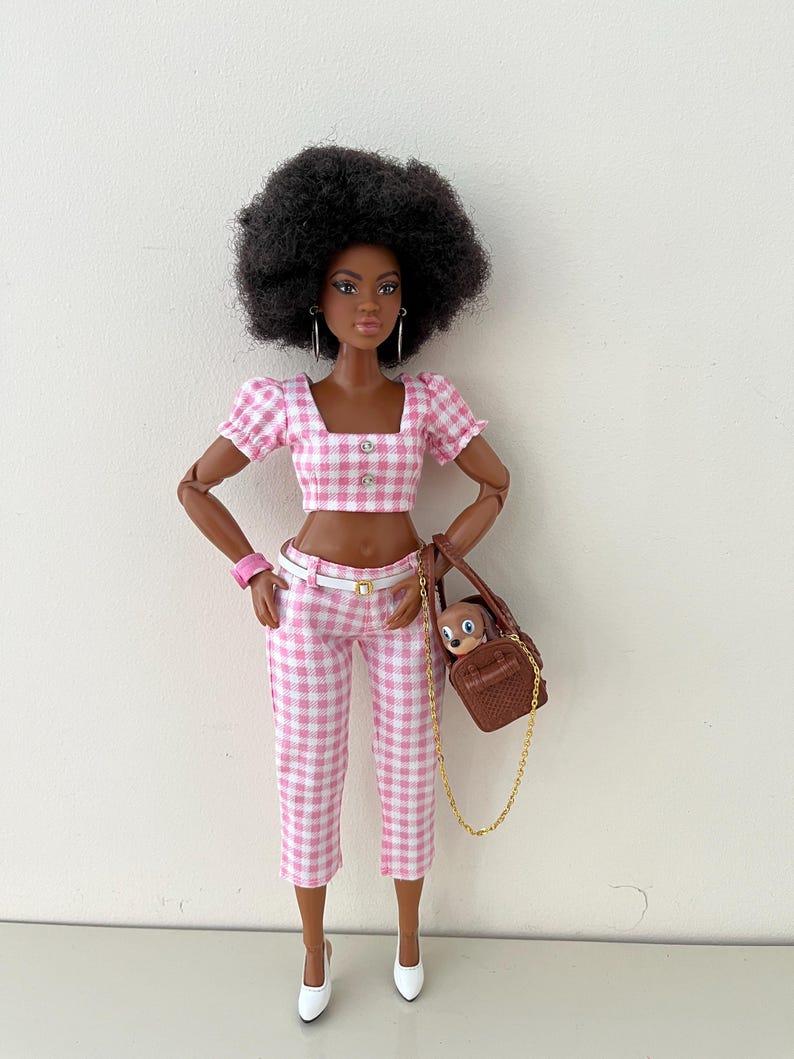 May include: A doll with dark skin and a black afro hairstyle wearing a pink and white gingham crop top with puff sleeves and matching cropped trousers. The doll has a white belt, silver hoop earrings, a pink wristband, white heels, and a brown handbag.