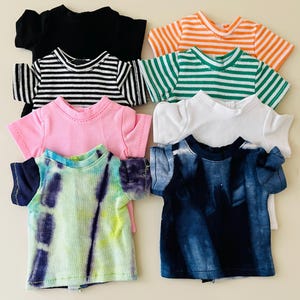 May include: A collection of seven small doll t-shirts in various patterns and colors, including solid black, white, pink, orange and white stripes, green and white stripes, black and white stripes, a blue tie-dye, and a green and purple tie-dye.