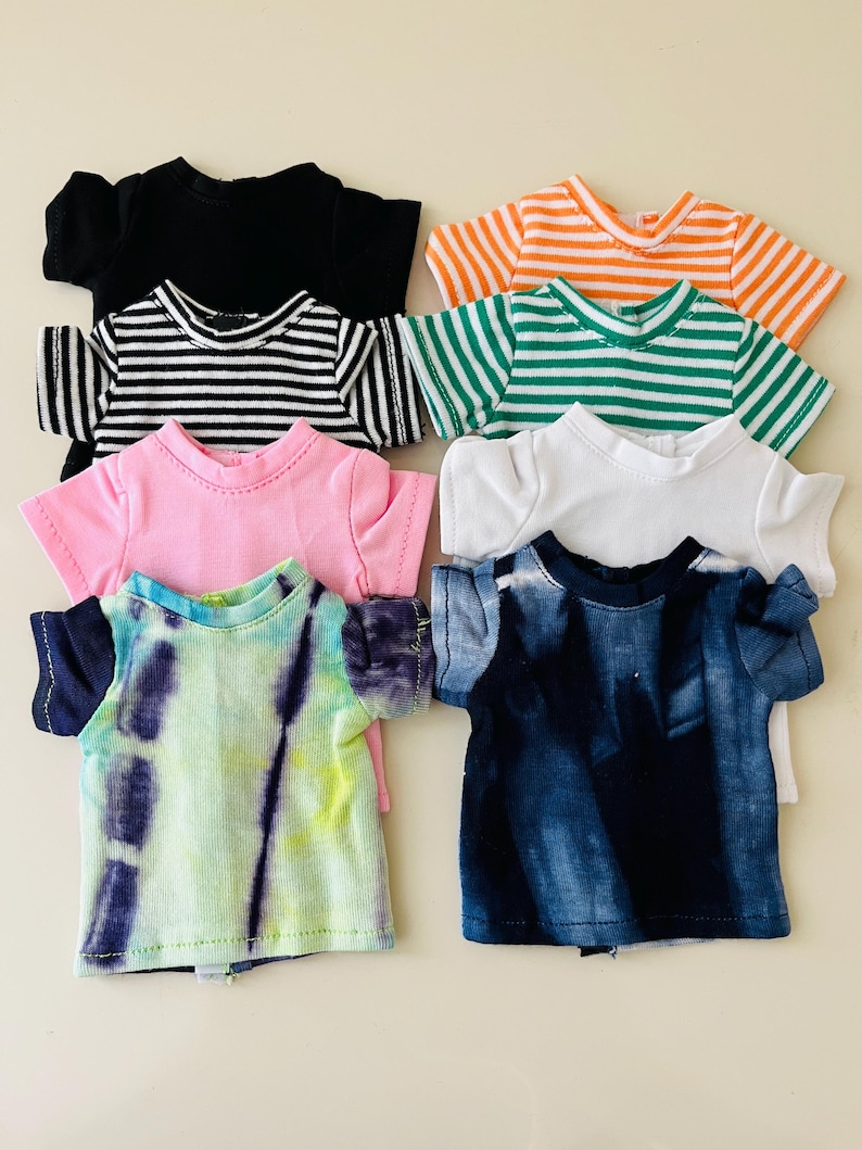 Puede incluir: Una colecci&oacute;n de siete camisetas peque&ntilde;as para mu&ntilde;ecas con diversos patrones y colores, incluyendo negro s&oacute;lido, blanco, rosa, rayas naranjas y blancas, rayas verdes y blancas, rayas negras y blancas, un tie-dye azul y un tie-dye verde y morado.