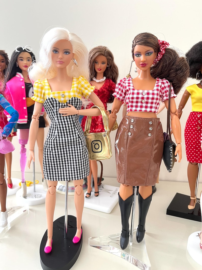 May include: Two fashion dolls standing side by side. The left doll has blonde hair, a yellow and white gingham puff-sleeve top, and a black and white gingham dress. The right doll has brown hair, a red and white gingham crop top, and a brown skirt.