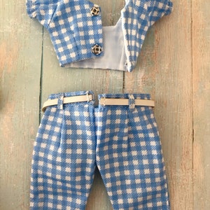 May include: A two-piece doll outfit featuring a light blue and white gingham pattern. The top is a short-sleeved cropped blouse with two silver metal snap closures. The matching trousers include a thin white belt threaded through belt loops.