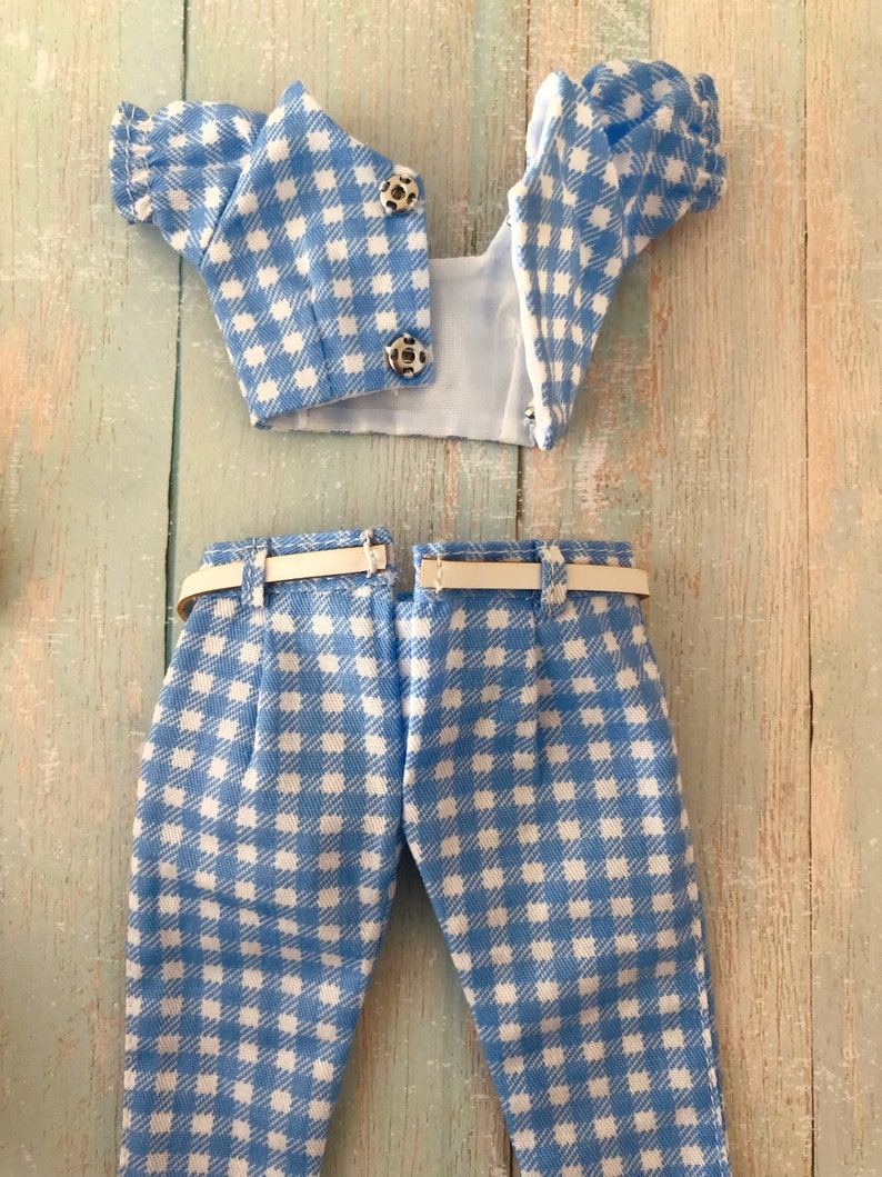 May include: A two-piece doll outfit featuring a light blue and white gingham pattern. The top is a short-sleeved cropped blouse with two silver metal snap closures. The matching trousers include a thin white belt threaded through belt loops.