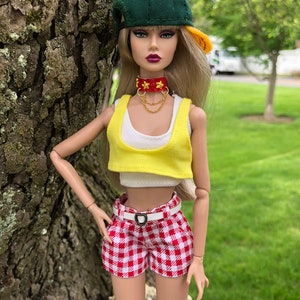 May include: A fashion doll with long blonde hair wearing a green and yellow baseball cap, a red choker with gold stars and chains, a white tank top, a yellow crop top, and red and white gingham shorts with a white belt featuring a heart buckle.