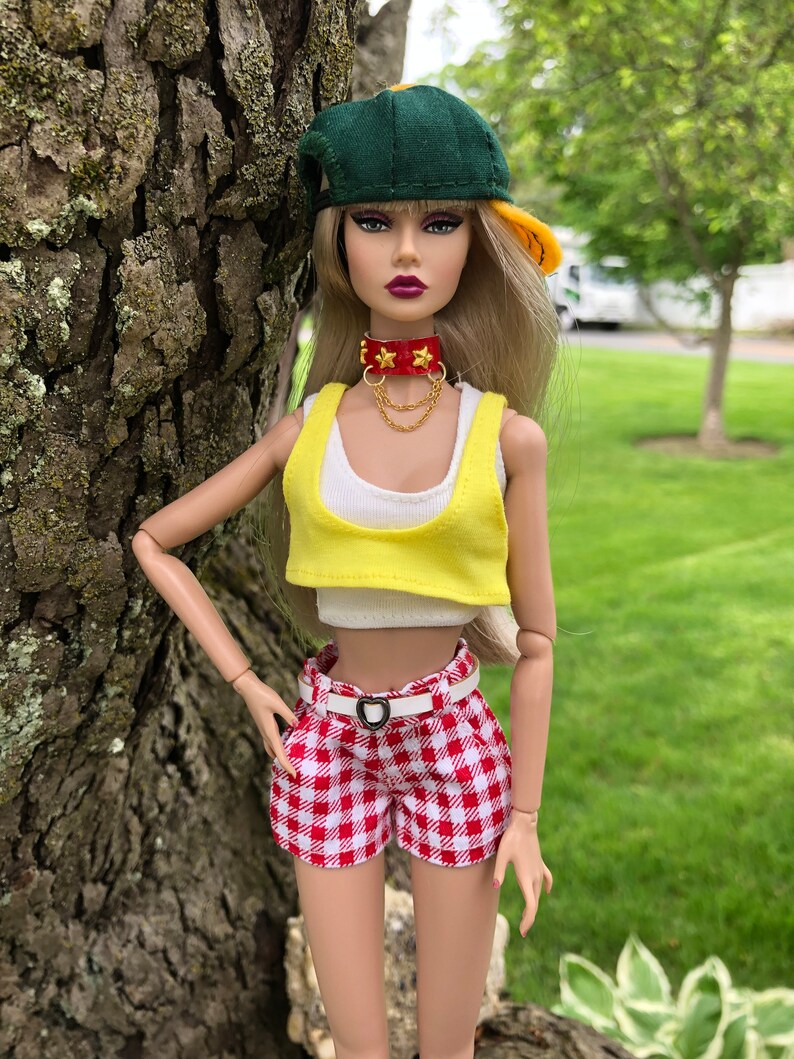 May include: A fashion doll with long blonde hair wearing a green and yellow baseball cap, a red choker with gold stars and chains, a white tank top, a yellow crop top, and red and white gingham shorts with a white belt featuring a heart buckle.