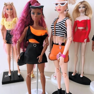 May include: Four fashion dolls standing on display stands. The dolls wear varied outfits including a yellow gingham top, a black and orange cut-out dress, a black and white striped crop top with an orange bag, and a red two-piece set with a skirt.
