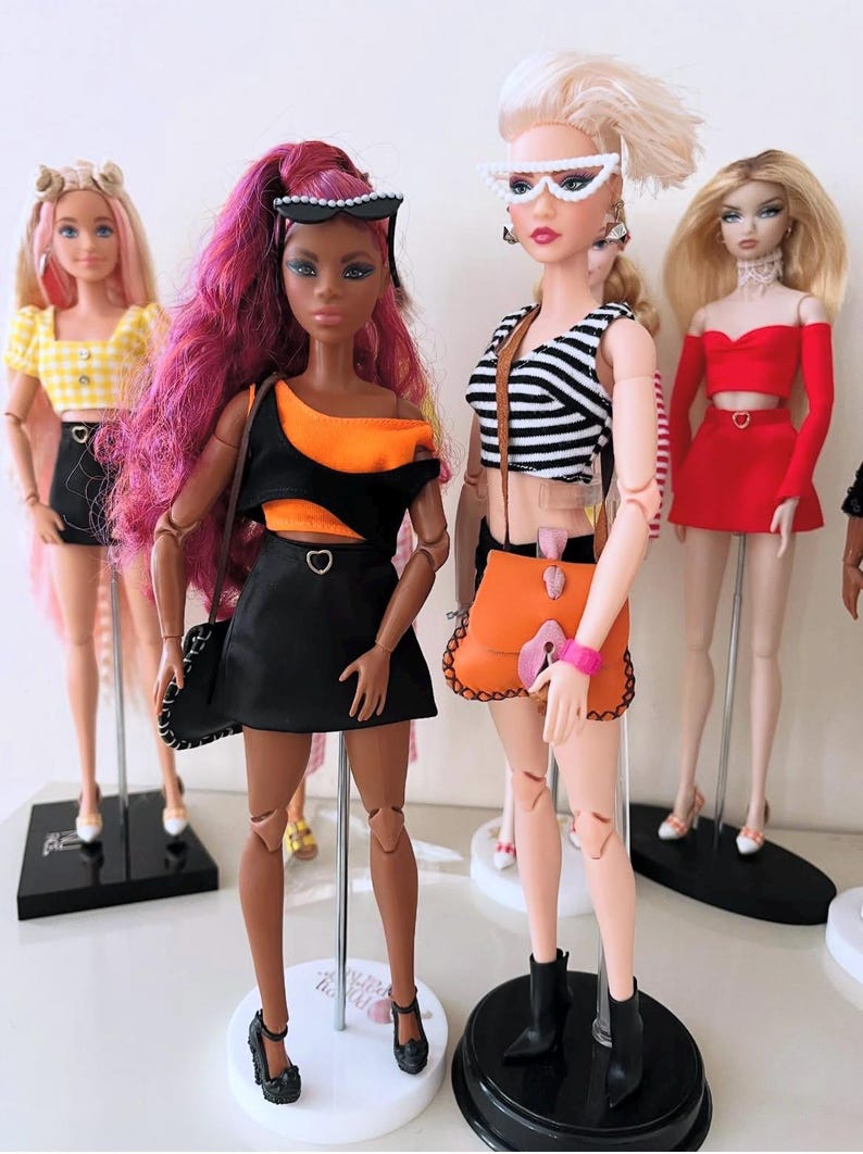 May include: Four fashion dolls standing on display stands. The dolls wear varied outfits including a yellow gingham top, a black and orange cut-out dress, a black and white striped crop top with an orange bag, and a red two-piece set with a skirt.