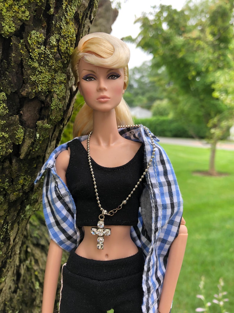 May include: A fashion doll with blonde hair styled in a pompadour, wearing a black crop top, black pants, and an unbuttoned blue, white, and black plaid shirt. A silver-toned ball chain necklace with a clear rhinestone cross pendant is worn.