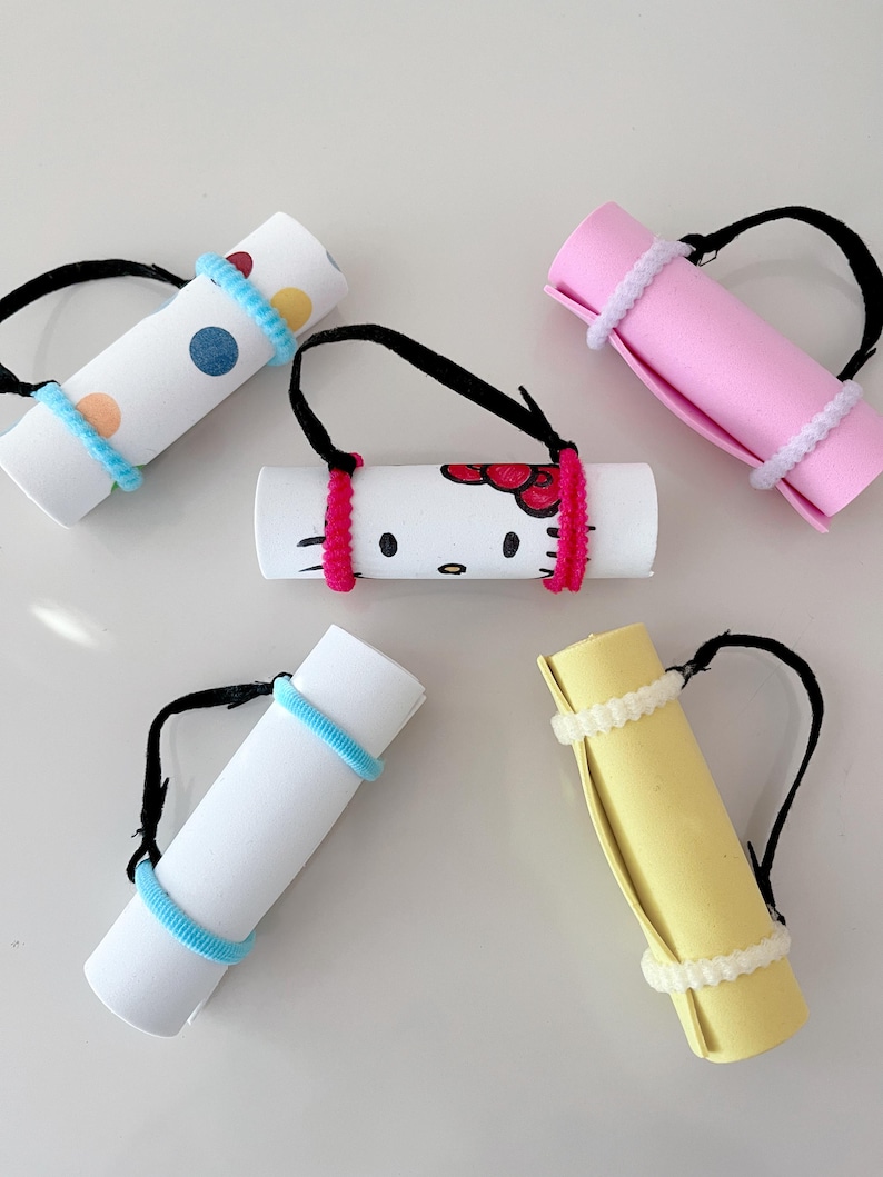 May include: Five colorful rolled-up mats with handles. The mats are decorated with polka dots, Hello Kitty, and solid colors. The handles are made of black elastic.