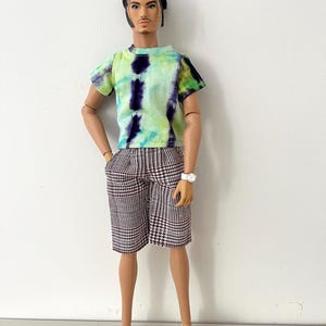 Puede incluir: Una mu&ntilde;eca de moda con cabello oscuro recogido en un mo&ntilde;o, que viste una camiseta de manga corta con estampado tie-dye en tonos verde lima y morado oscuro, combinada con pantalones cortos hasta la rodilla con patr&oacute;n de pata de gallo marr&oacute;n y blanco, un reloj blanco y mocasines blancos.