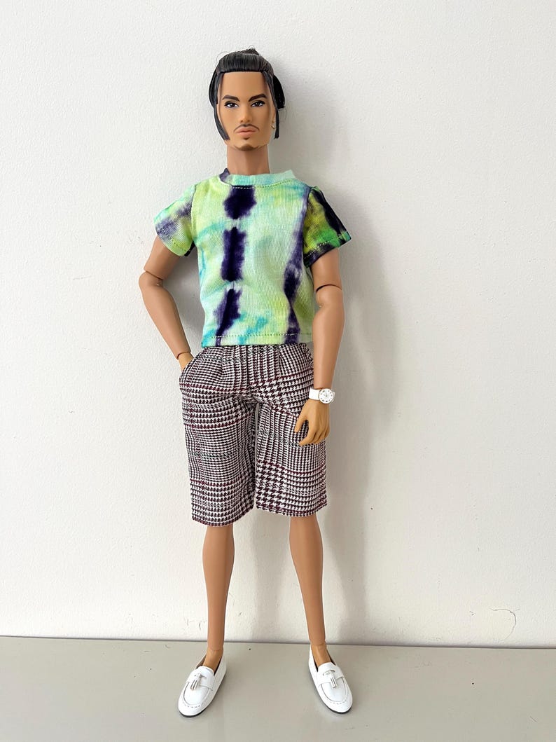 Puede incluir: Una mu&ntilde;eca de moda con cabello oscuro recogido en un mo&ntilde;o, que viste una camiseta de manga corta con estampado tie-dye en tonos verde lima y morado oscuro, combinada con pantalones cortos hasta la rodilla con patr&oacute;n de pata de gallo marr&oacute;n y blanco, un reloj blanco y mocasines blancos.