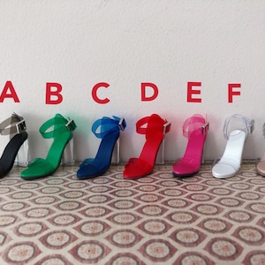 Op de afbeelding: Acht paar transparante sandalen met hak en verschillende gekleurde bandjes. De sandalen zijn in een rij van links naar rechts gerangschikt en van A tot en met G gelabeld. De sandalen hebben een transparante hak en een transparant bandje dat om de enkel gaat. De bandjes zijn zwart, groen, blauw, rood, roze, wit en beige.