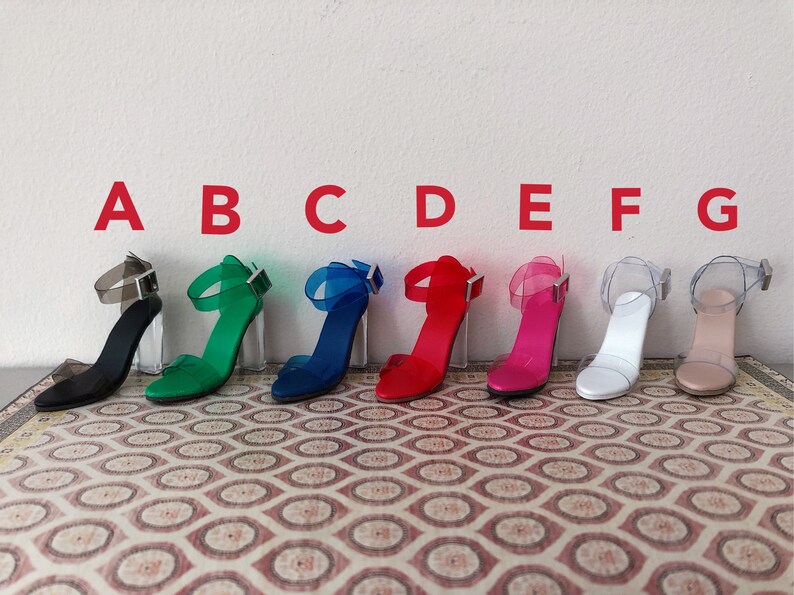 Op de afbeelding: Acht paar transparante sandalen met hak en verschillende gekleurde bandjes. De sandalen zijn in een rij van links naar rechts gerangschikt en van A tot en met G gelabeld. De sandalen hebben een transparante hak en een transparant bandje dat om de enkel gaat. De bandjes zijn zwart, groen, blauw, rood, roze, wit en beige.
