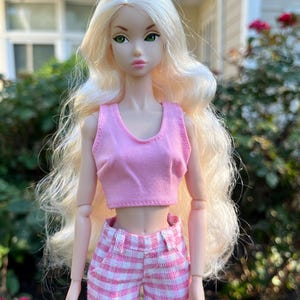 May include: A doll with long wavy blonde hair and green eyes wearing a light pink sleeveless crop top and pink and white checkered shorts. The doll has articulated joints at the shoulders and elbows and a neutral facial expression with pink lips.