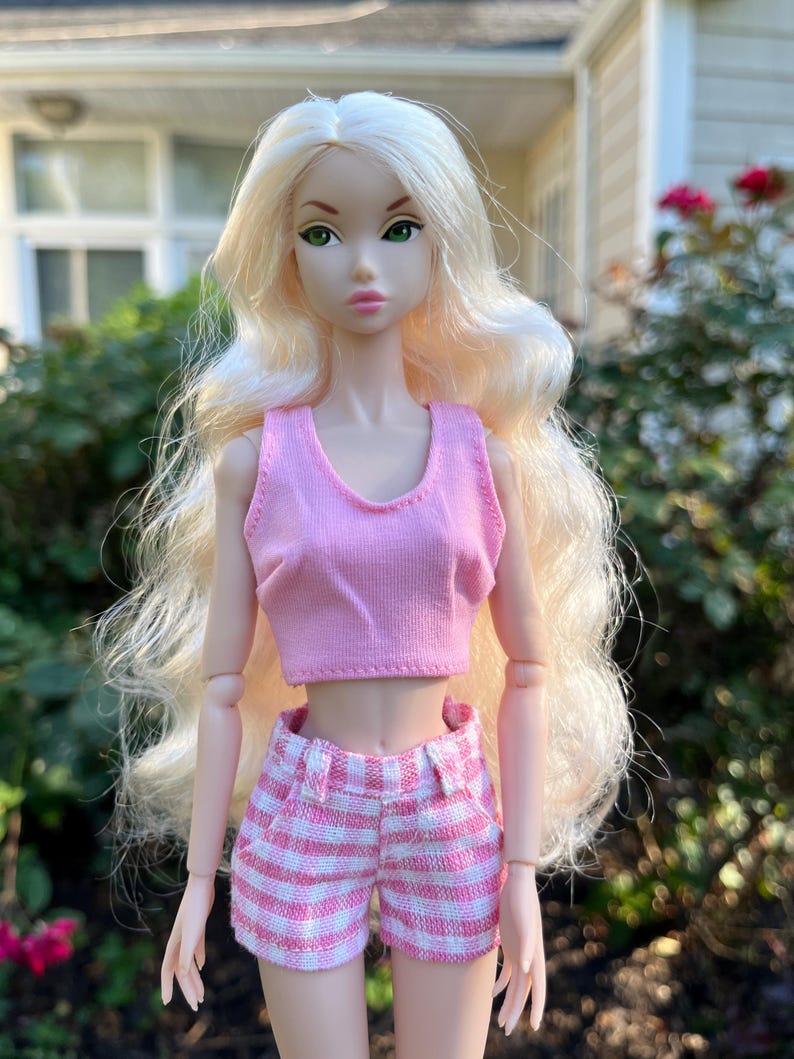 May include: A doll with long wavy blonde hair and green eyes wearing a light pink sleeveless crop top and pink and white checkered shorts. The doll has articulated joints at the shoulders and elbows and a neutral facial expression with pink lips.