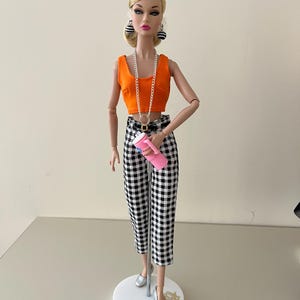 May include: A fashion doll with blonde hair styled in a low bun, wearing an orange sleeveless crop top, black and white gingham capri pants, silver flat shoes, black and white spherical earrings, a long silver chain necklace, and holding a pink tumbler.