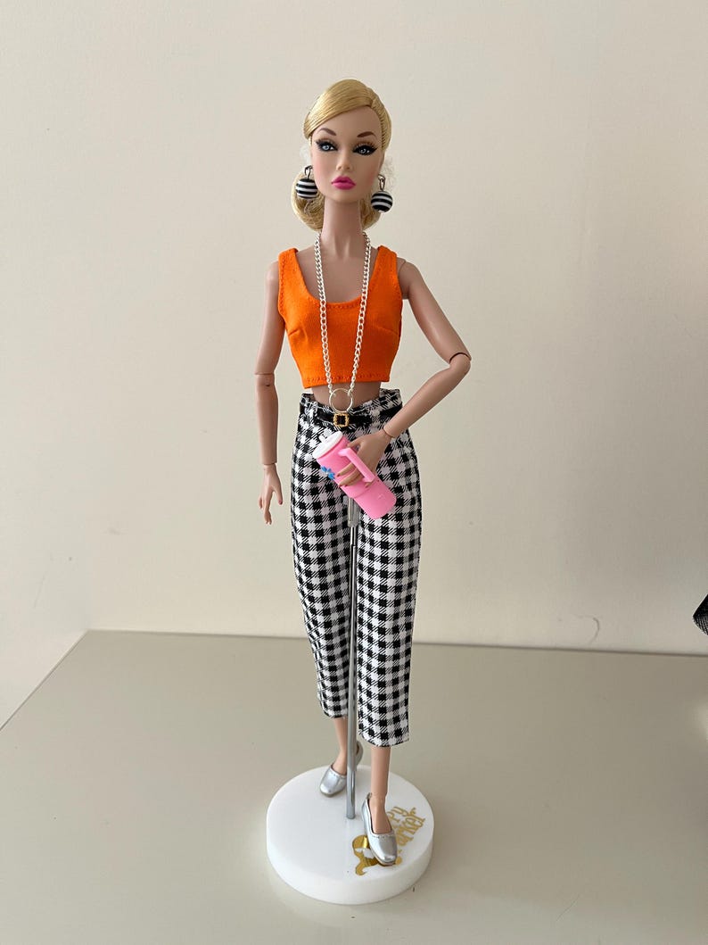 May include: A fashion doll with blonde hair styled in a low bun, wearing an orange sleeveless crop top, black and white gingham capri pants, silver flat shoes, black and white spherical earrings, a long silver chain necklace, and holding a pink tumbler.