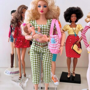 May include: A blonde doll wearing a green and white gingham crop top and matching capri pants, holding a small baby doll. The doll carries a pink and white checkered tote bag containing a blue bottle, standing among other dolls in various outfits.
