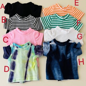Puede incluir: Una colecci&oacute;n de ocho camisetas de mu&ntilde;eca de manga corta etiquetadas de la A a la H. Las camisetas incluyen una negra s&oacute;lida, una a rayas blancas y negras, una rosa, una tie-dye verde y morada, una a rayas naranjas y blancas, una a rayas verdes y blancas, una blanca y una tie-dye azul.