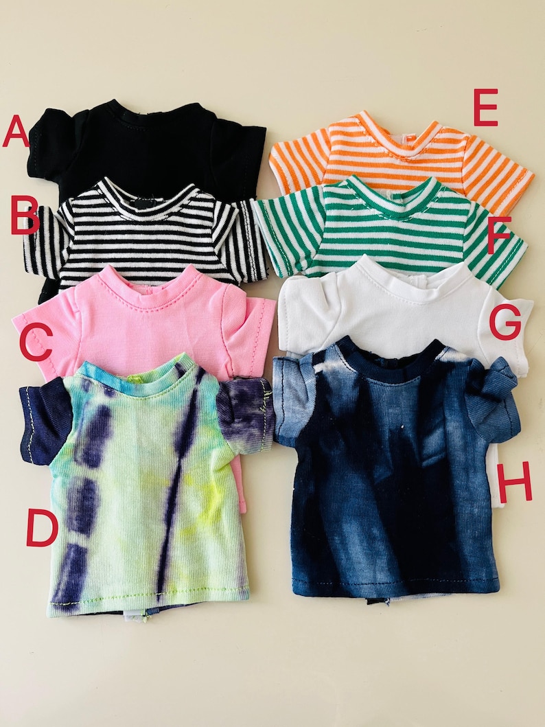 Puede incluir: Una colecci&oacute;n de ocho camisetas de mu&ntilde;eca de manga corta etiquetadas de la A a la H. Las camisetas incluyen una negra s&oacute;lida, una a rayas blancas y negras, una rosa, una tie-dye verde y morada, una a rayas naranjas y blancas, una a rayas verdes y blancas, una blanca y una tie-dye azul.