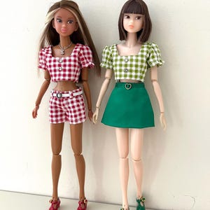 May include: Two fashion dolls standing side by side. The left doll wears a red and white gingham crop top and matching shorts with a white belt. The right doll wears a green and white gingham crop top and a solid green skirt with a heart-shaped belt.