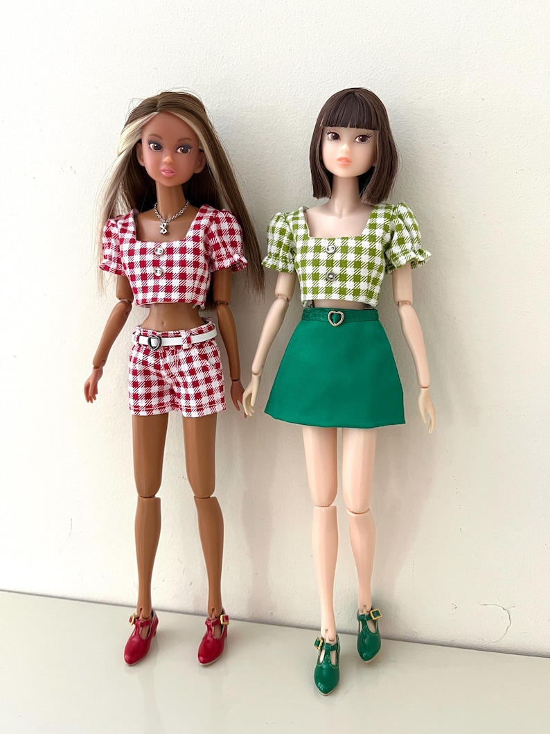 May include: Two fashion dolls standing side by side. The left doll wears a red and white gingham crop top and matching shorts with a white belt. The right doll wears a green and white gingham crop top and a solid green skirt with a heart-shaped belt.